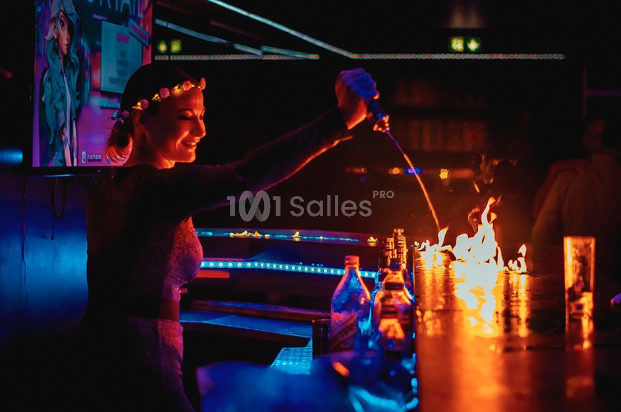Une femme verse un liquide enflammé sur un bar dans une ambiance sombre et lumineuse de club.