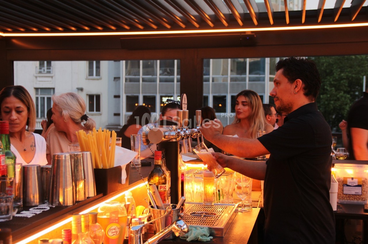 Un barman sert des boissons dans un bar animé avec des clients assis autour du comptoir éclairé.