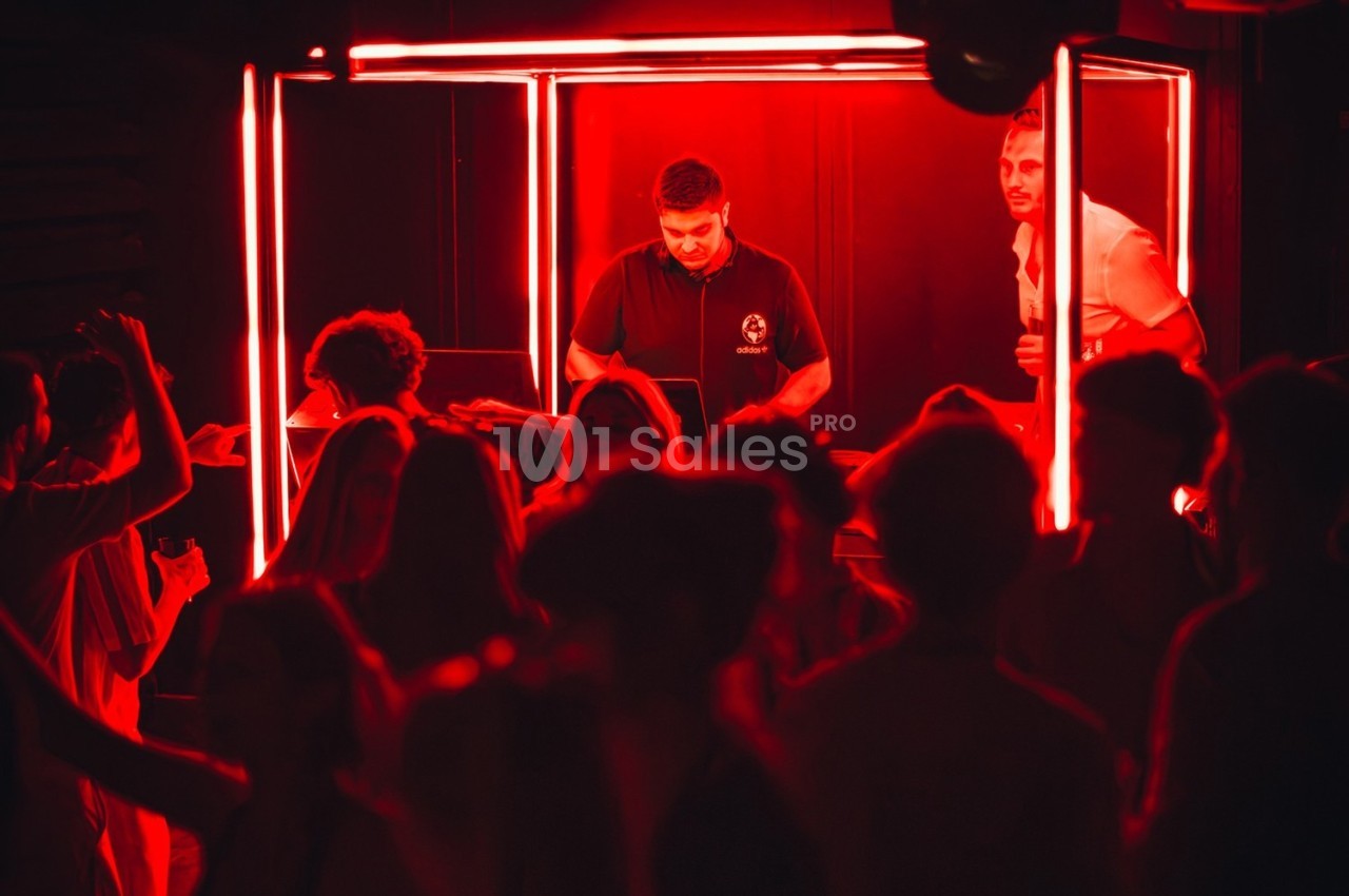 Un DJ mixe dans une cabine éclairée de rouge, entouré de personnes dansant dans une ambiance sombre.