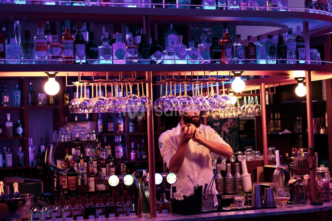 Un barman prépare un cocktail derrière un comptoir éclairé, entouré de bouteilles et de verres suspendus.