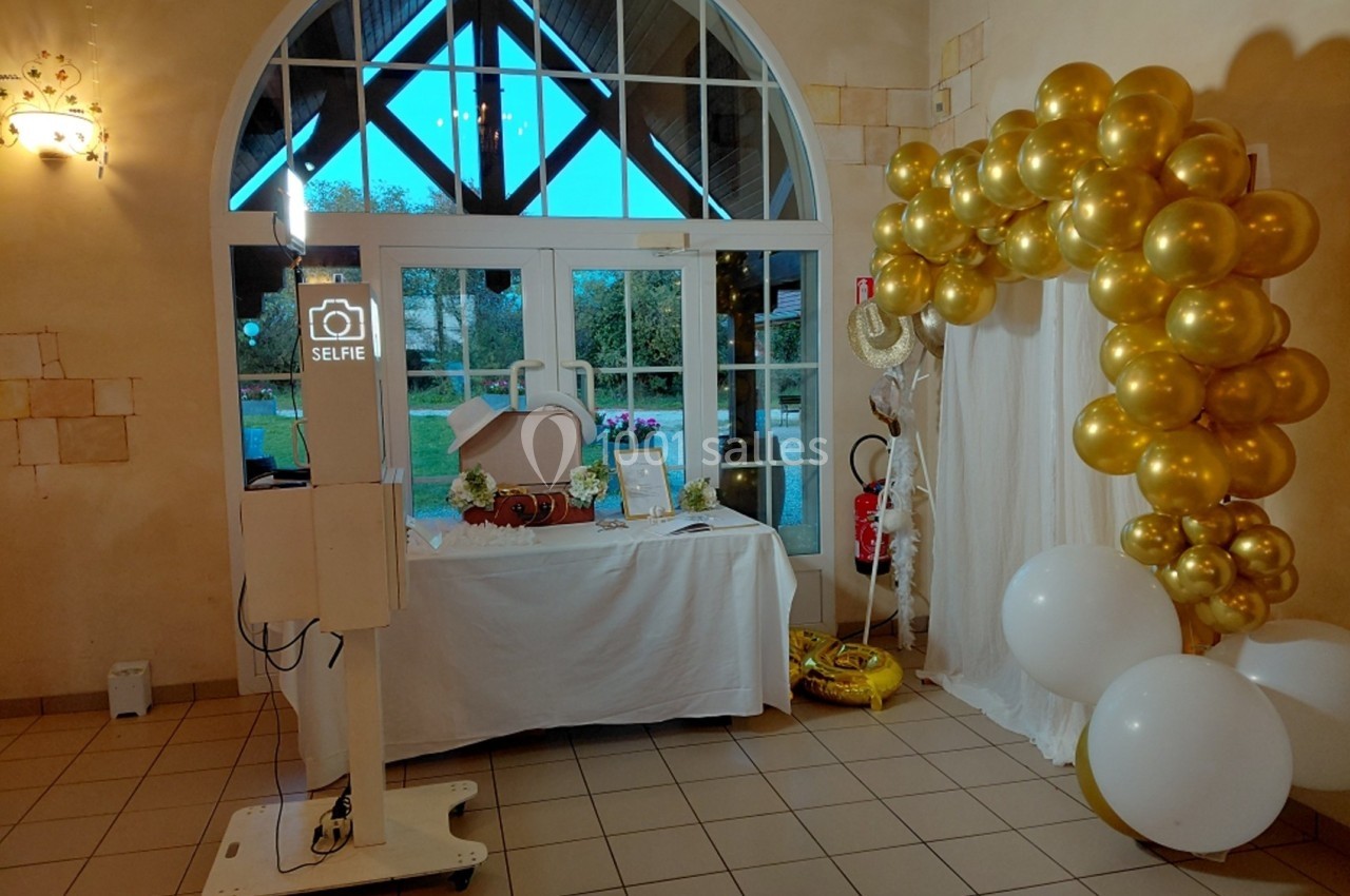 Borne photo avec décor de ballons dorés et table ornée pour un événement dans une salle lumineuse.