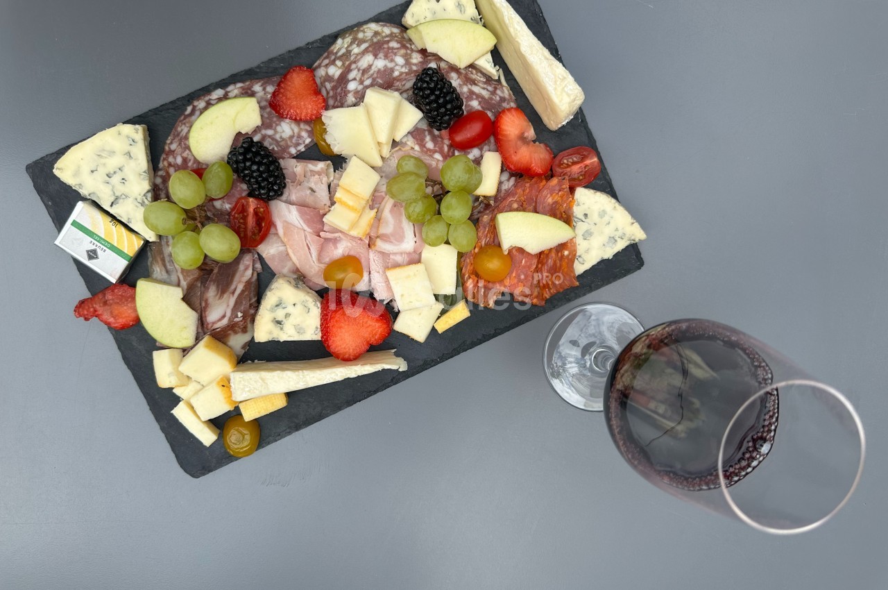 Plateau de charcuterie et fromages variés avec fruits frais, accompagné d'un verre de vin rouge sur une table grise.