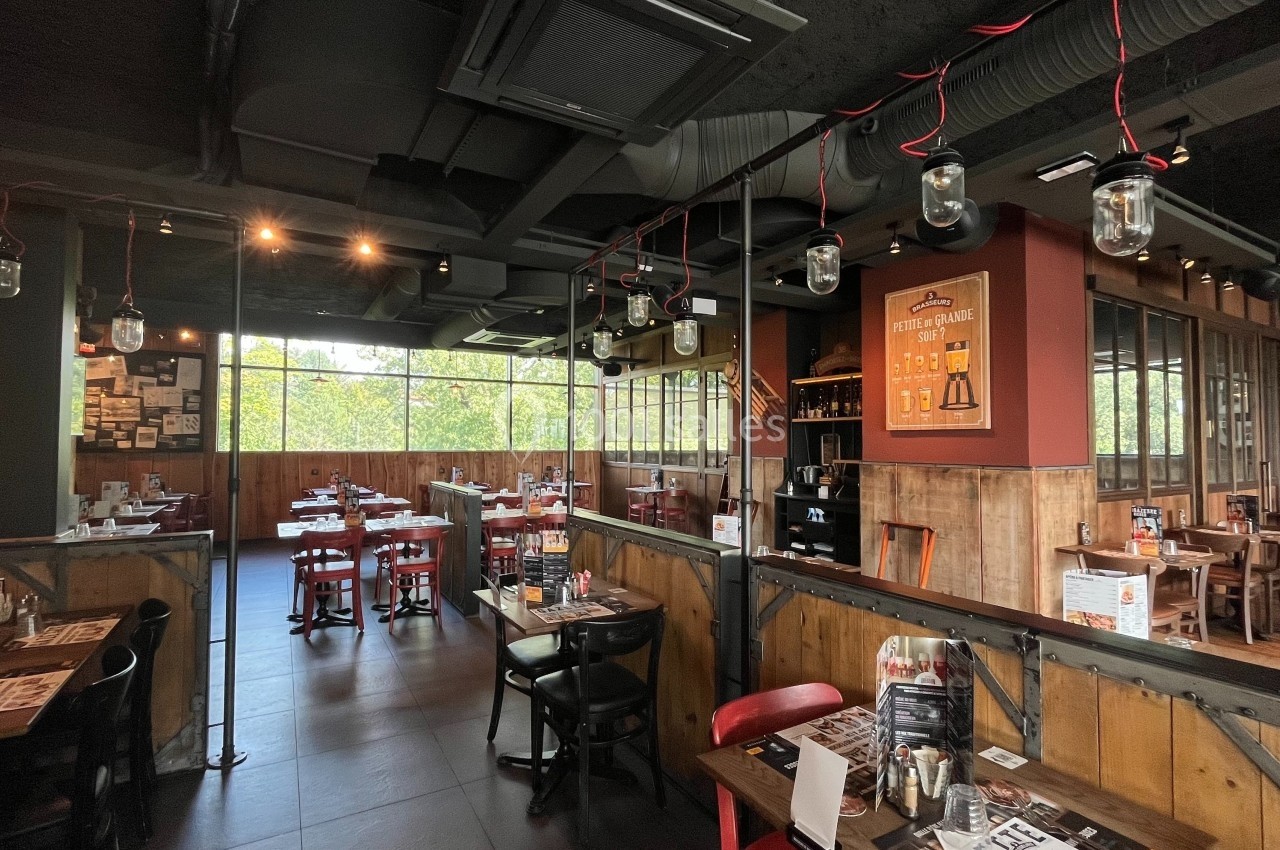 Salle de restaurant lumineuse avec tables en bois, chaises rouges et noires, décor industriel et grandes fenêtres.