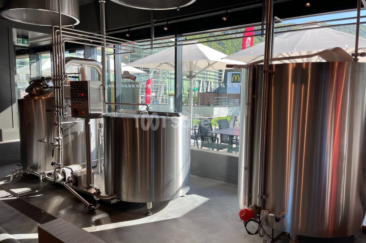 Cuves en acier inoxydable dans une brasserie, avec vue sur une terrasse extérieure et des parasols.