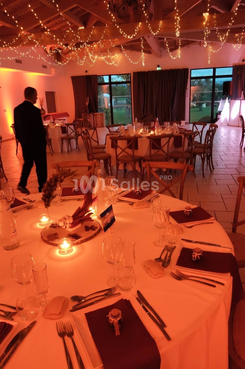 Salle de réception décorée avec des guirlandes lumineuses, tables dressées avec nappes blanches et serviettes bordeaux.
