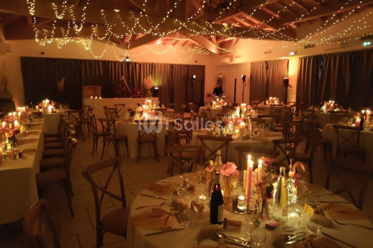 Salle de réception décorée pour un dîner, avec tables dressées, bougies allumées et guirlandes lumineuses suspendues.
