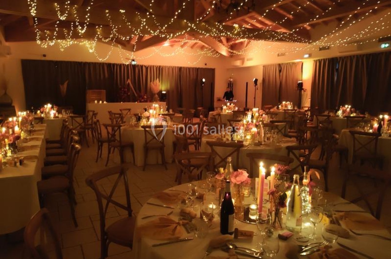 Salle de réception décorée pour un mariage, avec tables dressées, bougies et guirlandes lumineuses suspendues.