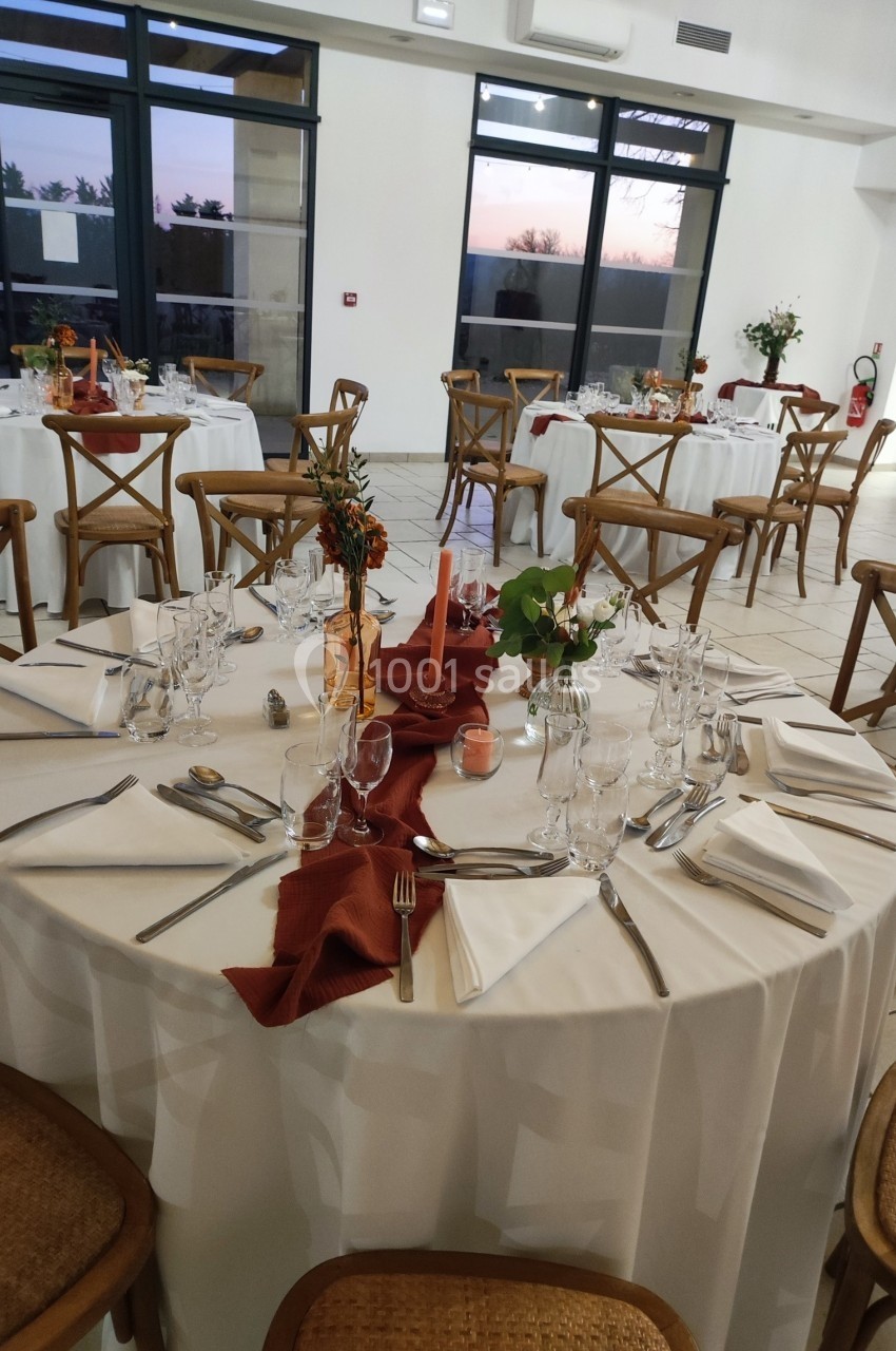 Salle de réception avec tables rondes dressées, nappes blanches, serviettes rouges et chaises en bois.