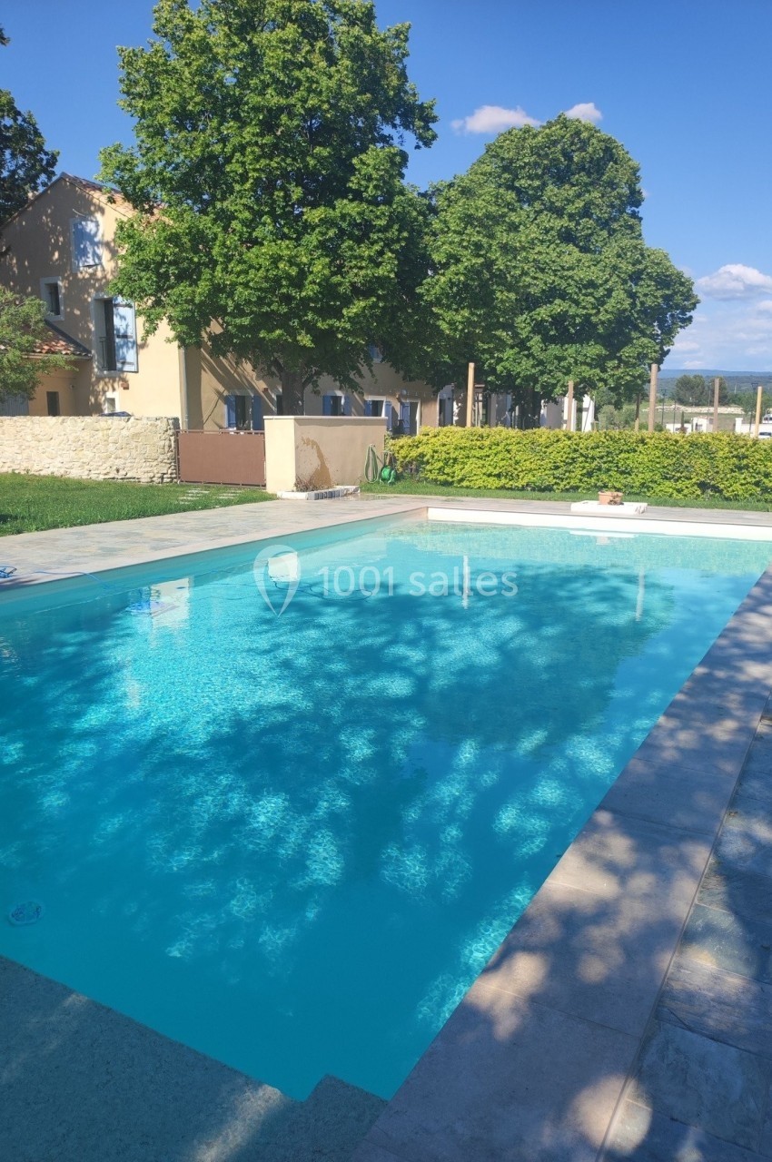 Piscine extérieure entourée de verdure et de bâtiments en pierre sous un ciel bleu dégagé.