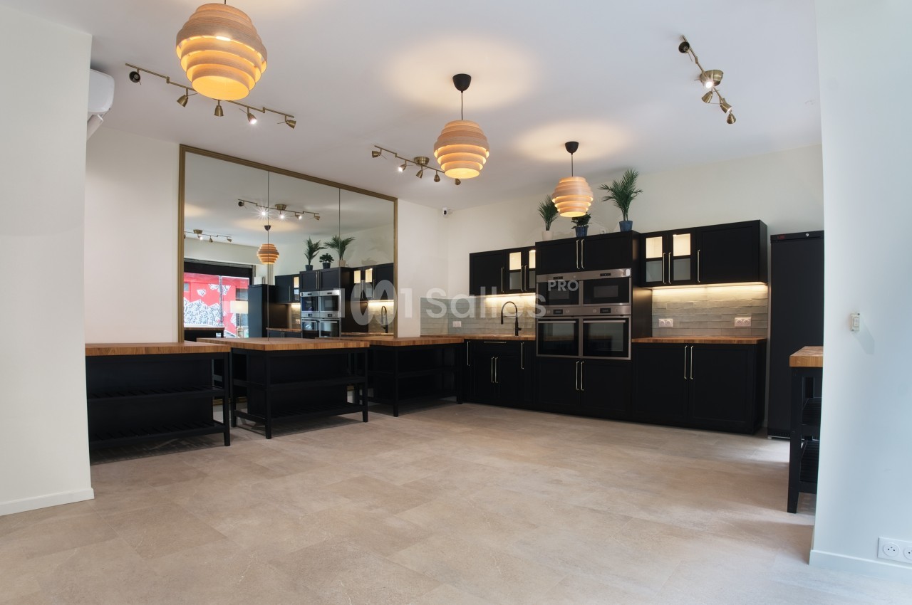 Cuisine moderne avec meubles noirs, plan de travail en bois, éclairage suspendu et grand miroir mural.