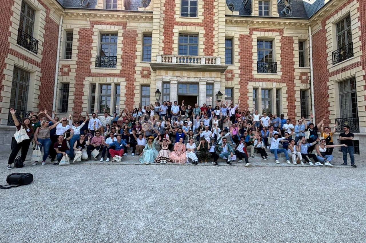 Groupe de personnes posant devant un grand bâtiment en briques rouges et pierres claires, par une journée ensoleillée.
