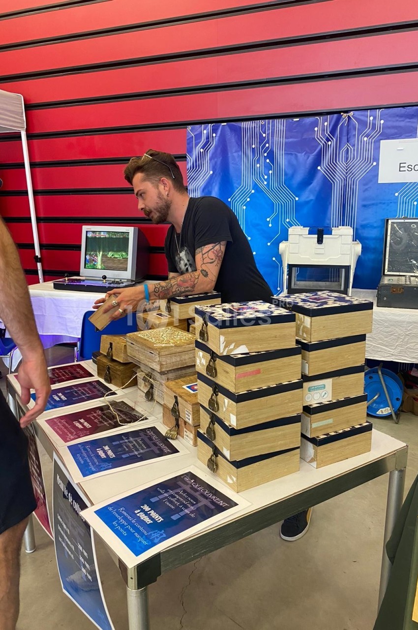 Un homme présente des boîtes en bois et des affiches sur une table lors d'un événement dans un espace intérieur.