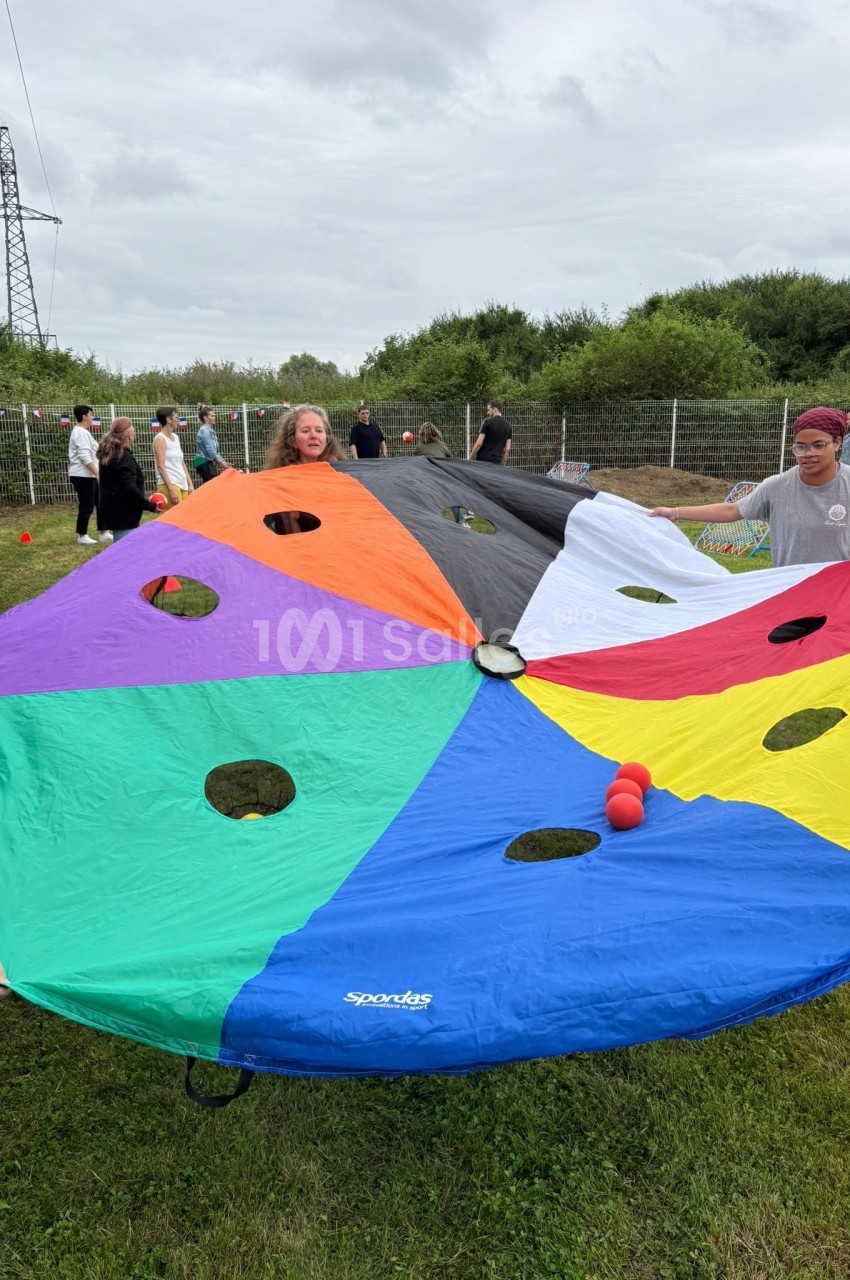 Des personnes tiennent un parachute coloré avec des trous, utilisé pour une activité de groupe en extérieur sur une pelouse.