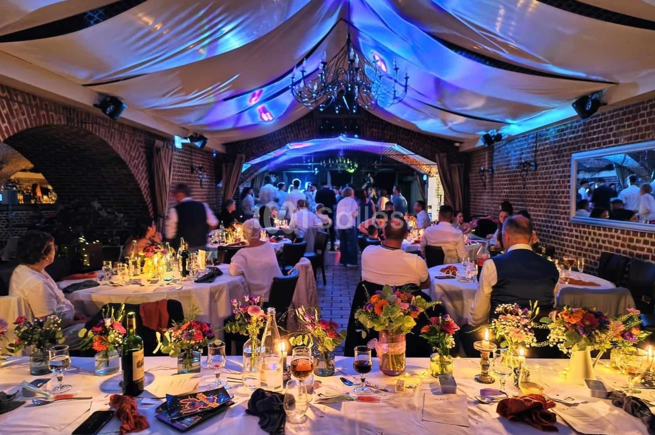 Salle de réception décorée avec des tables fleuries, des invités assis et une piste de danse éclairée en arrière-plan.