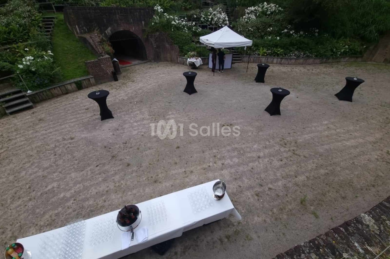 Espace extérieur aménagé pour un événement avec tables hautes, buffet et tente blanche, entouré de verdure.