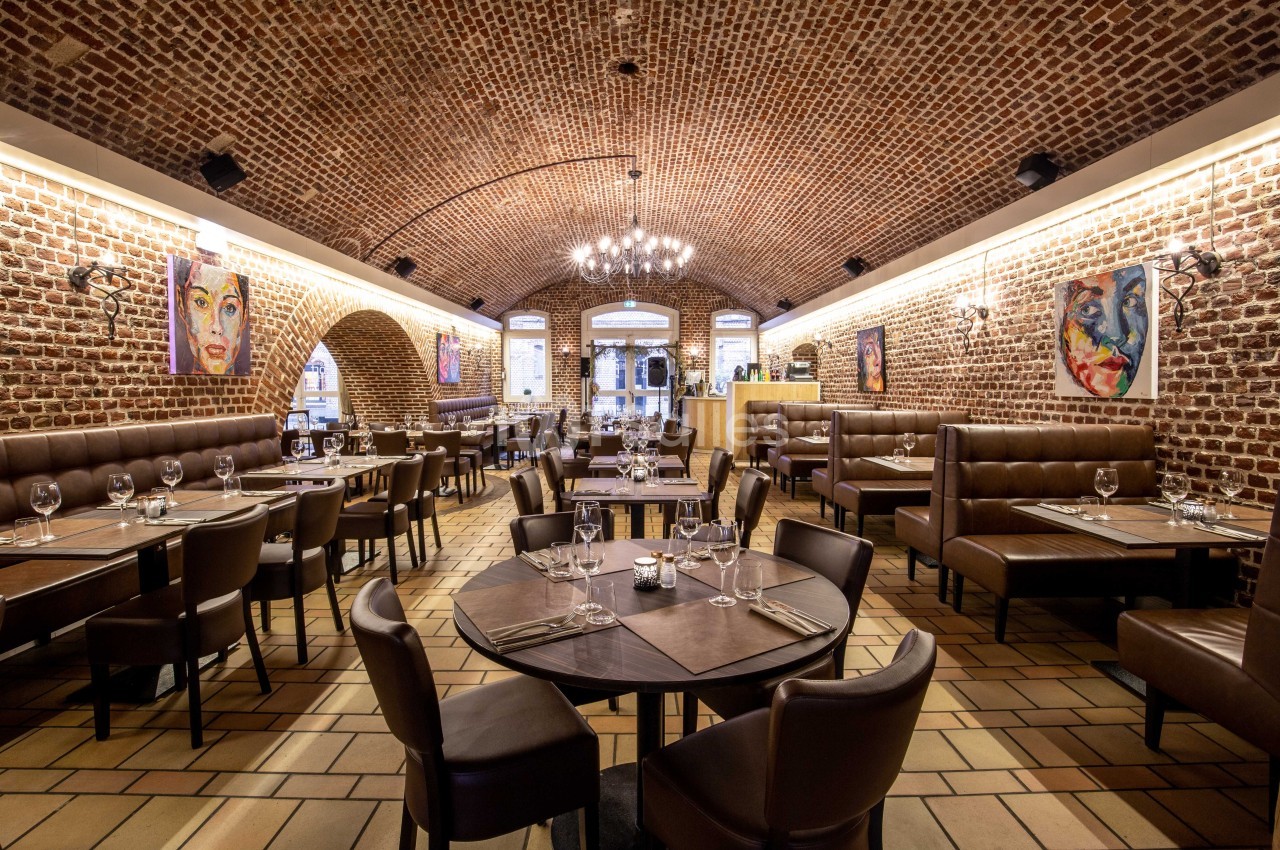 Salle de restaurant avec murs en briques, plafond voûté, tables dressées et peintures colorées accrochées aux murs.