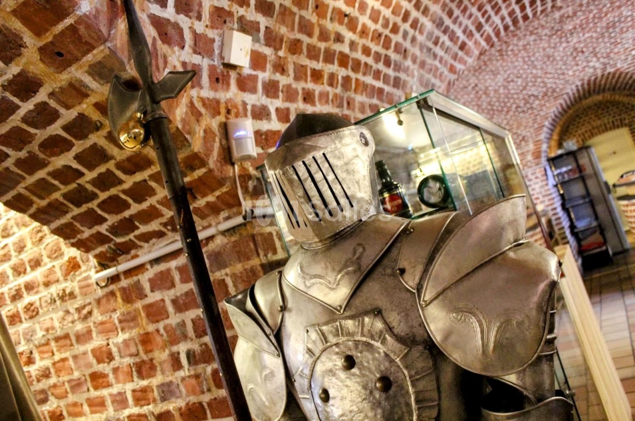 Armure médiévale en métal exposée dans une pièce voûtée en briques avec une vitrine en arrière-plan.