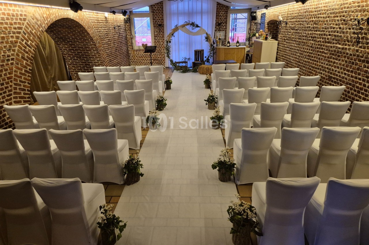 Salle de cérémonie décorée avec des chaises blanches alignées, une arche fleurie et des murs en briques apparentes.