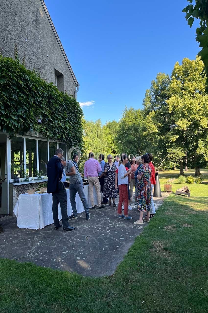 Groupe de personnes discutant debout près d'une table de buffet en extérieur, devant un bâtiment entouré de verdure.