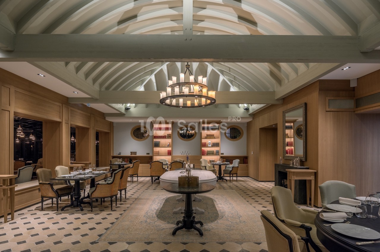 Salle de restaurant élégante avec poutres apparentes, lustre central, tables dressées et ambiance chaleureuse.