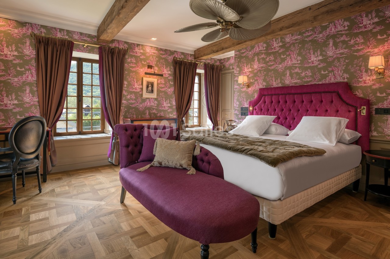 Chambre élégante avec lit double, canapé violet, parquet en bois et fenêtres encadrées de rideaux.