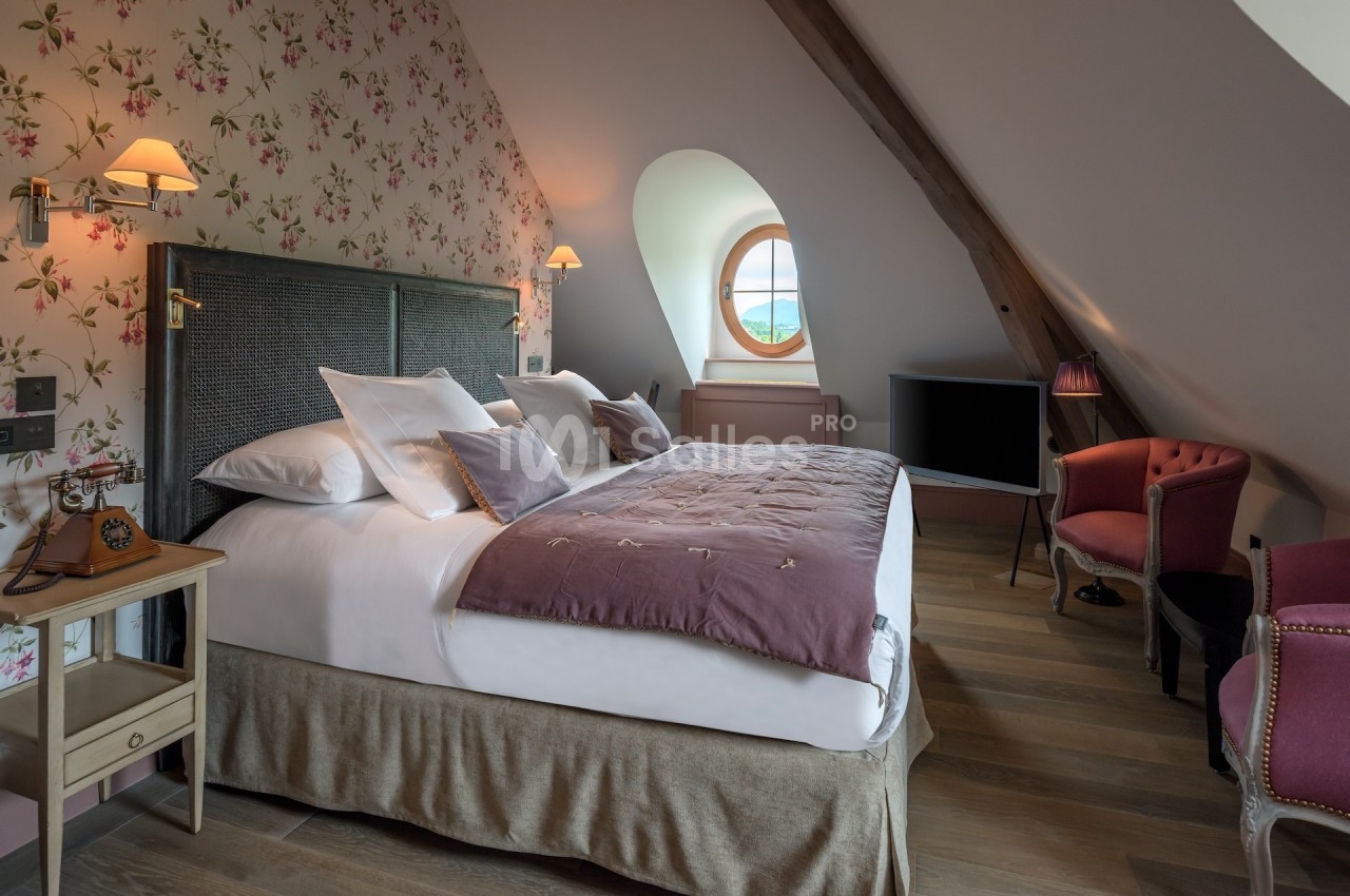 Chambre mansardée avec lit double, murs fleuris, fauteuils roses, fenêtre ronde et éclairage doux.