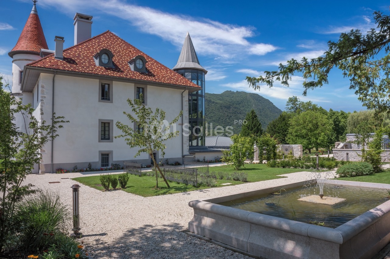 Manoir avec toit en tuiles rouges, jardin paysager, fontaine et montagnes en arrière-plan sous un ciel bleu.