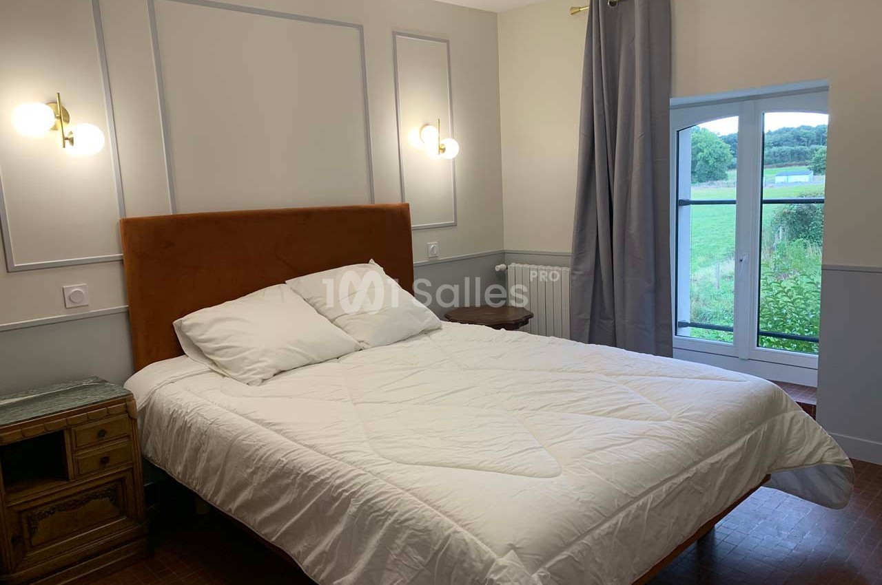Chambre lumineuse avec lit double, chevets en bois, murs clairs, rideaux gris et fenêtre donnant sur un paysage verdoyant.