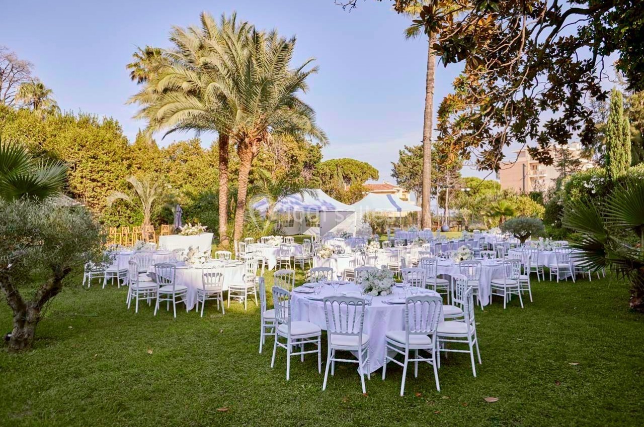 Tables rondes dressées avec nappes blanches disposées dans un jardin verdoyant entouré de palmiers et d'arbres.