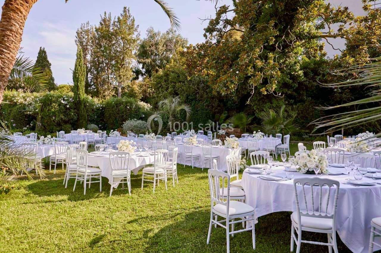 Tables rondes décorées de nappes blanches et de bouquets fleuris disposées dans un jardin verdoyant.