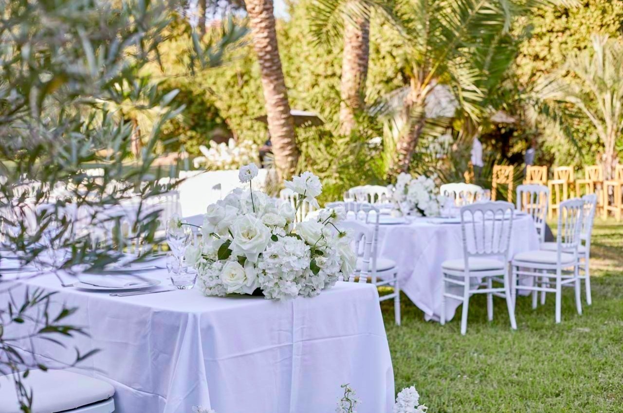 Tables décorées de fleurs blanches installées dans un jardin verdoyant avec des palmiers en arrière-plan.