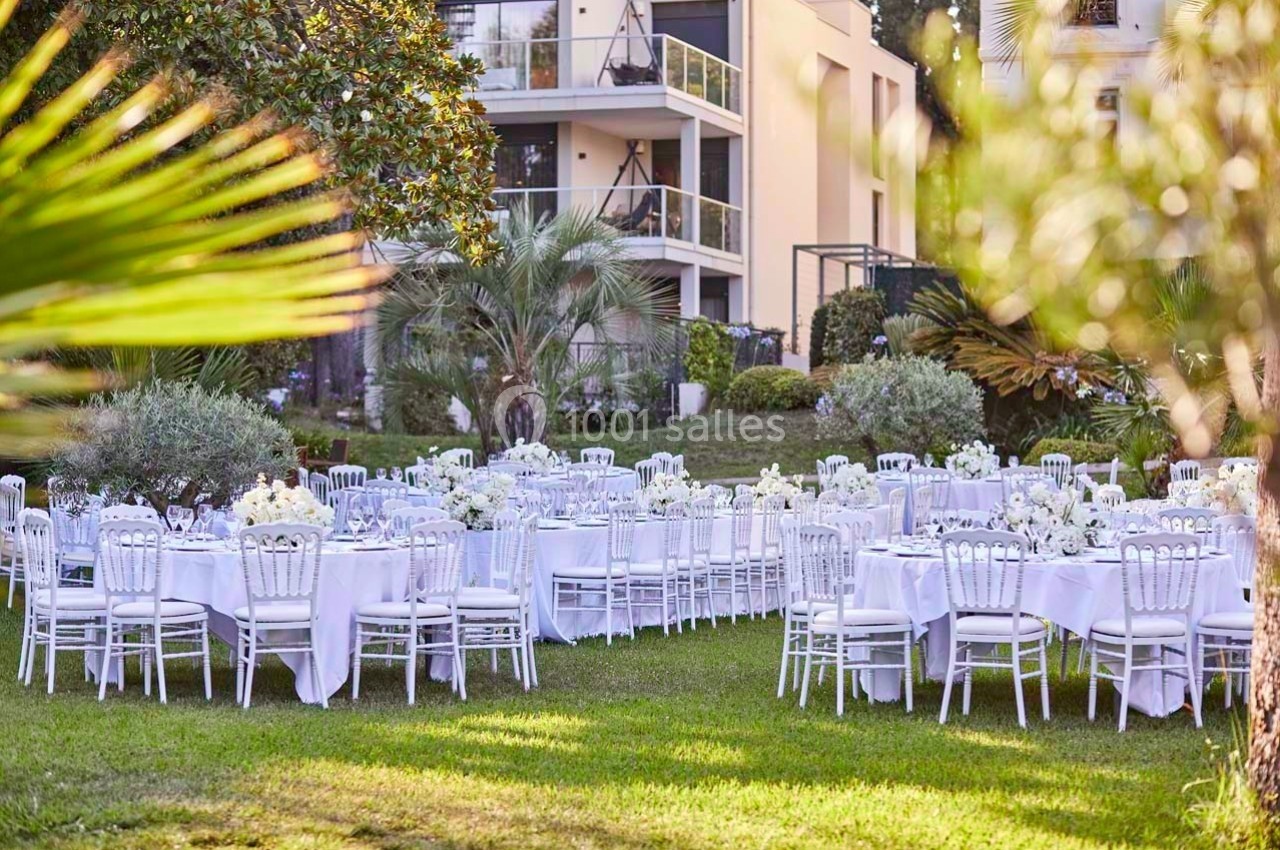 Tables rondes dressées avec nappes blanches et chaises élégantes dans un jardin verdoyant près d'un bâtiment moderne.