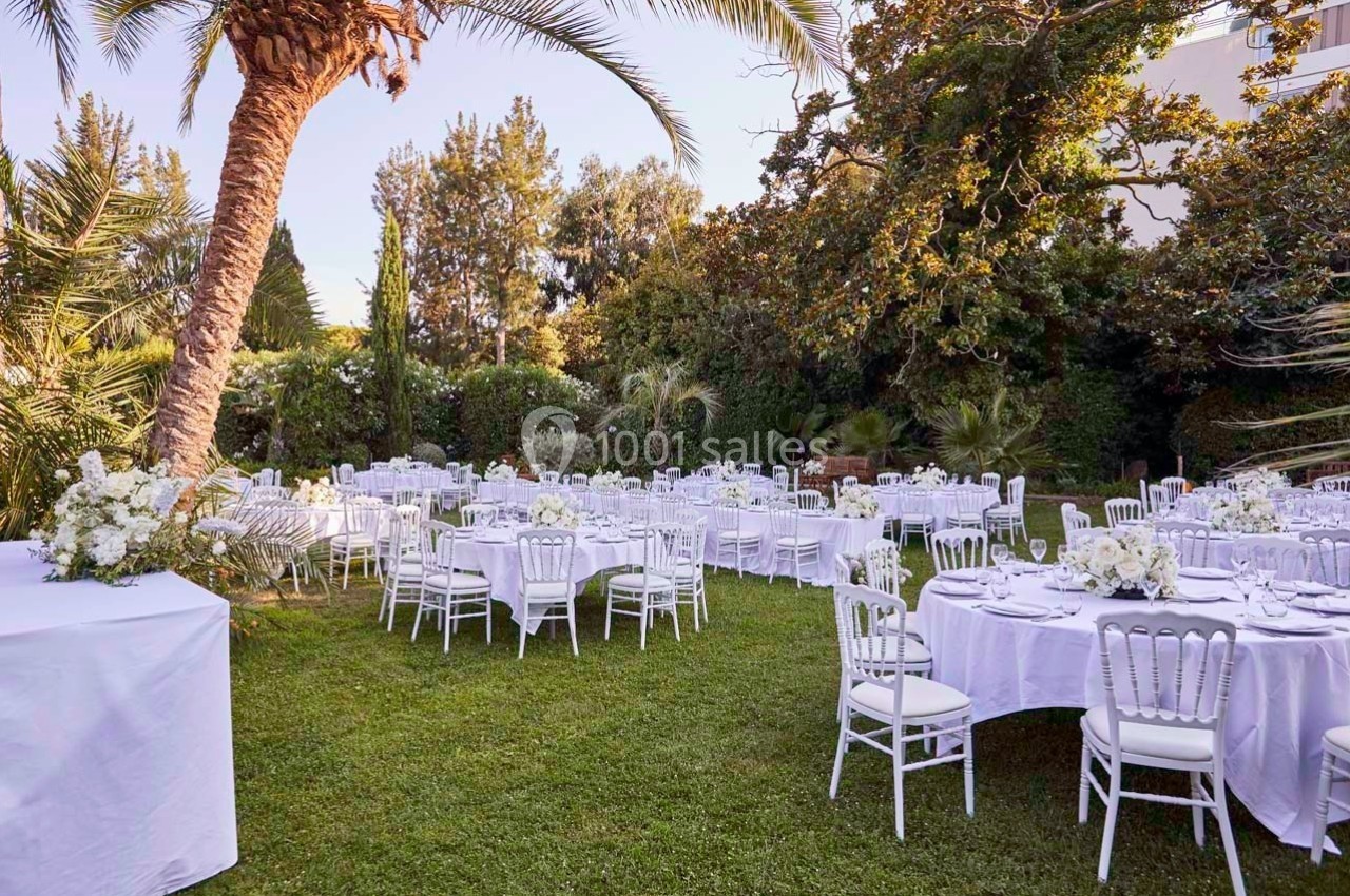 Tables rondes dressées avec nappes blanches et chaises blanches dans un jardin verdoyant sous des palmiers.