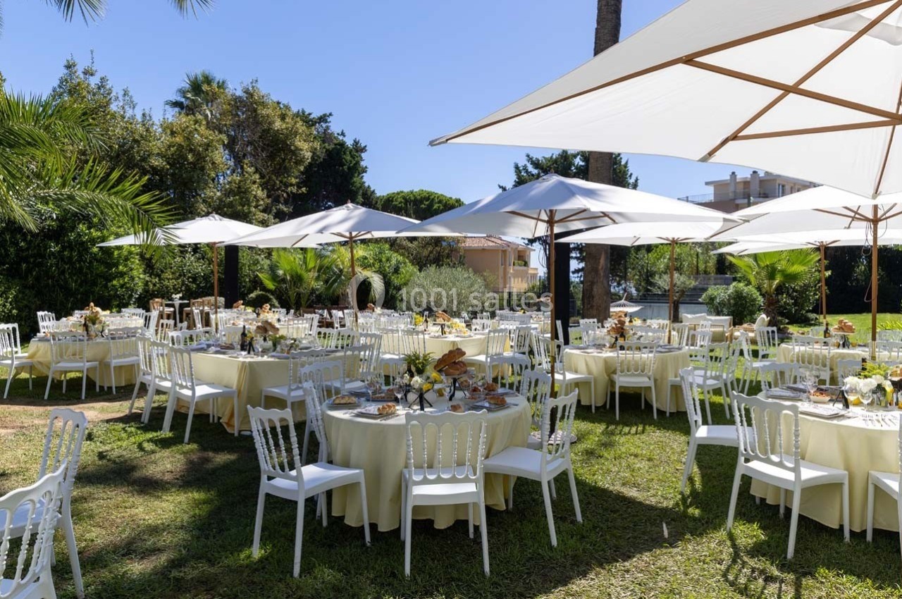 Tables rondes dressées avec nappes blanches et chaises, disposées sous des parasols dans un jardin ensoleillé.