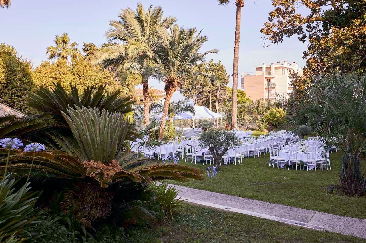 Jardin avec des tables et chaises blanches disposées pour un événement, entouré de palmiers et de végétation luxuriante.