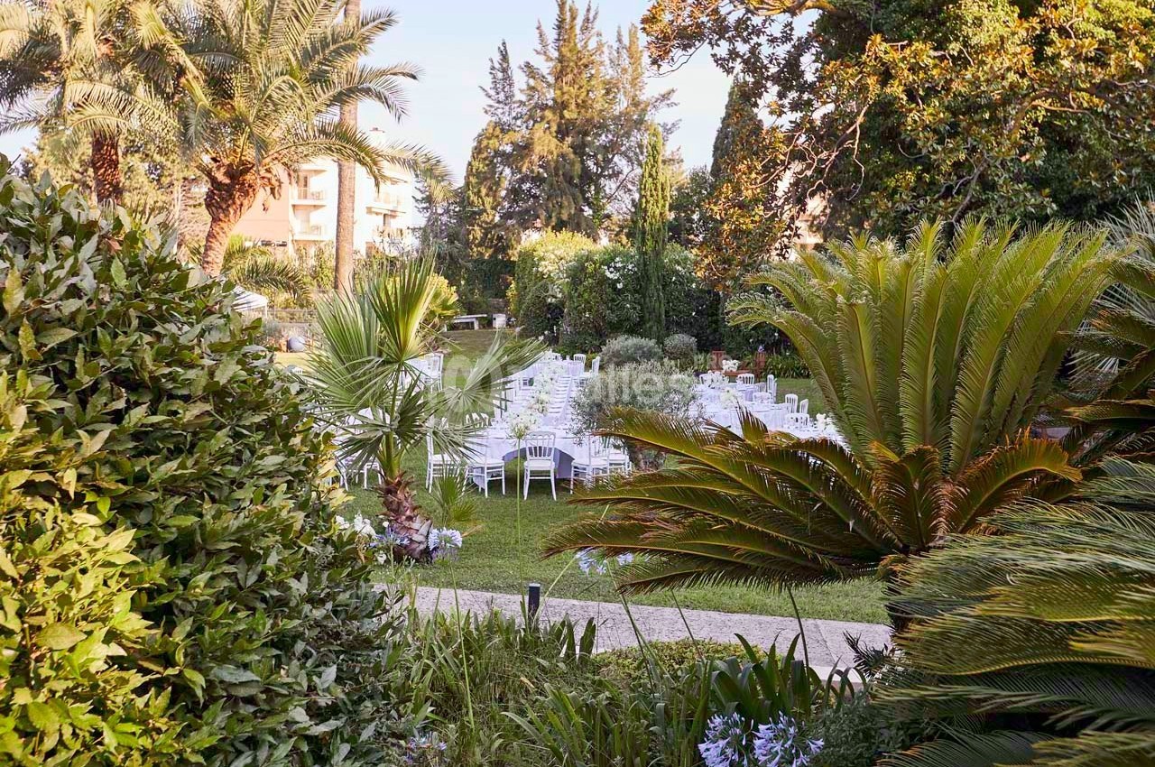 Jardin aménagé avec des tables et chaises blanches disposées pour un événement en plein air, entouré de végétation…