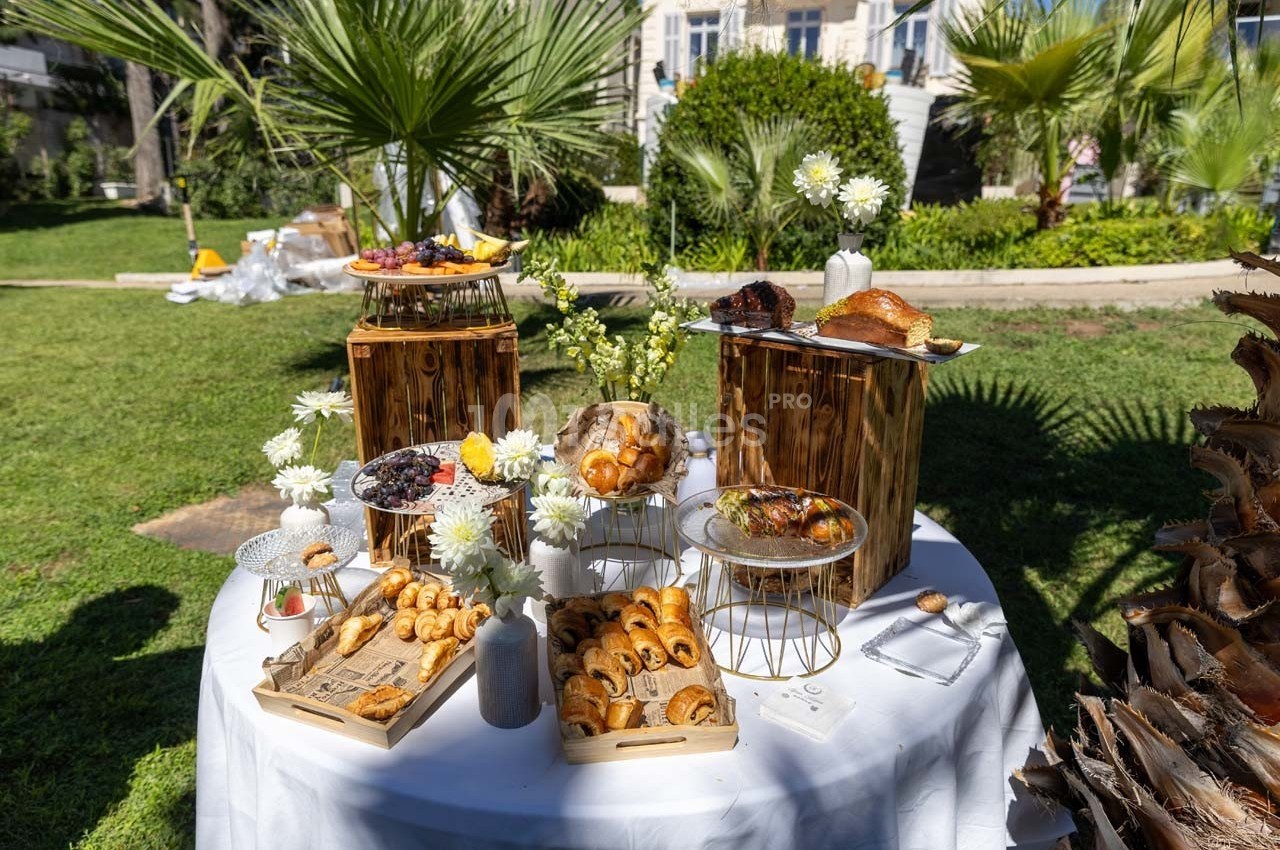 Table dressée en extérieur avec viennoiseries, pâtisseries et fruits, entourée de plantes et d'un jardin en arrière-plan.