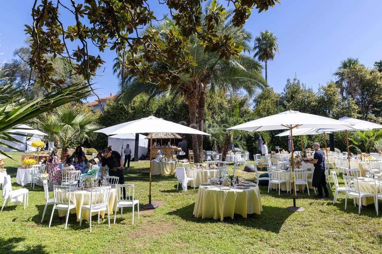 Jardin aménagé pour un événement avec tables rondes dressées, chaises blanches et parasols sous des arbres.
