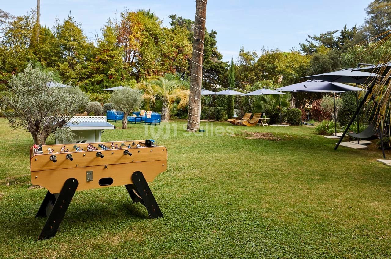 Table de baby-foot en bois placée sur une pelouse, entourée d'arbres, de parasols et de mobilier de jardin.