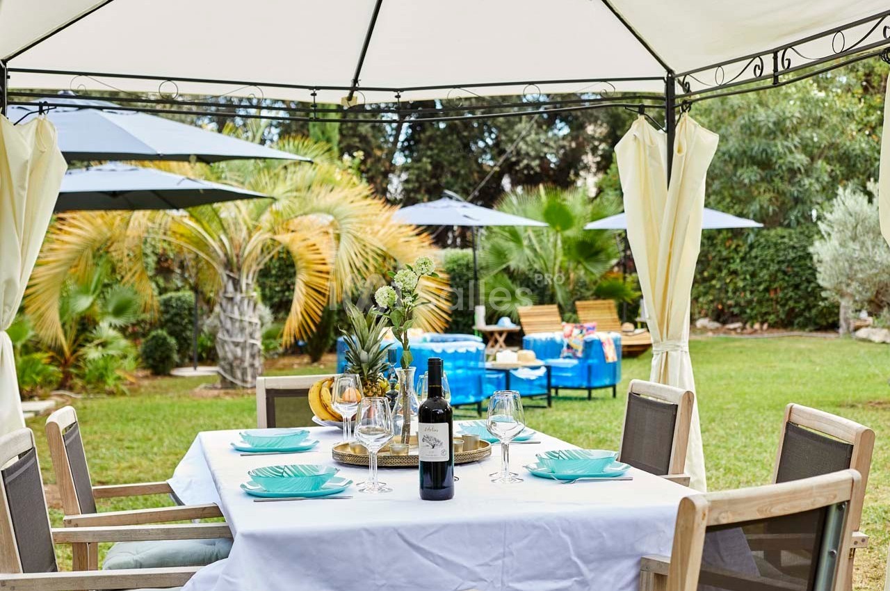 Table dressée sous une tonnelle dans un jardin avec chaises, vaisselle, bouteille de vin et vue sur des palmiers.