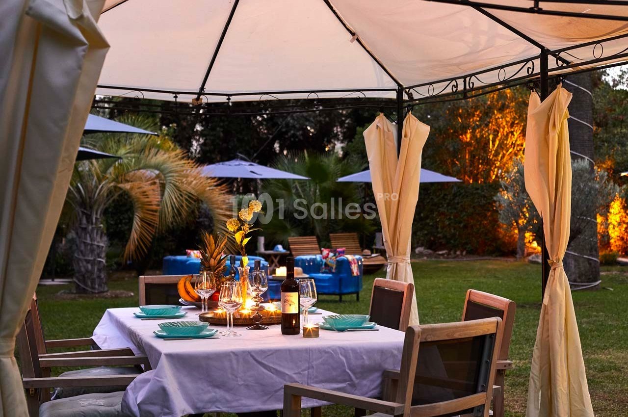 Table dressée sous une tonnelle dans un jardin, avec des bougies allumées et des chaises en bois autour.