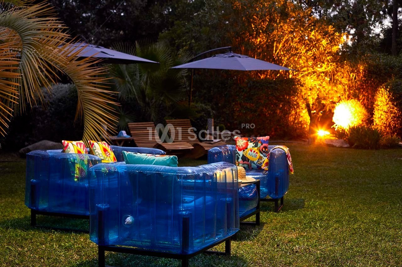 Mobilier de jardin coloré avec fauteuils bleus et coussins, éclairé par des lampes et un feu dans un espace extérieur…