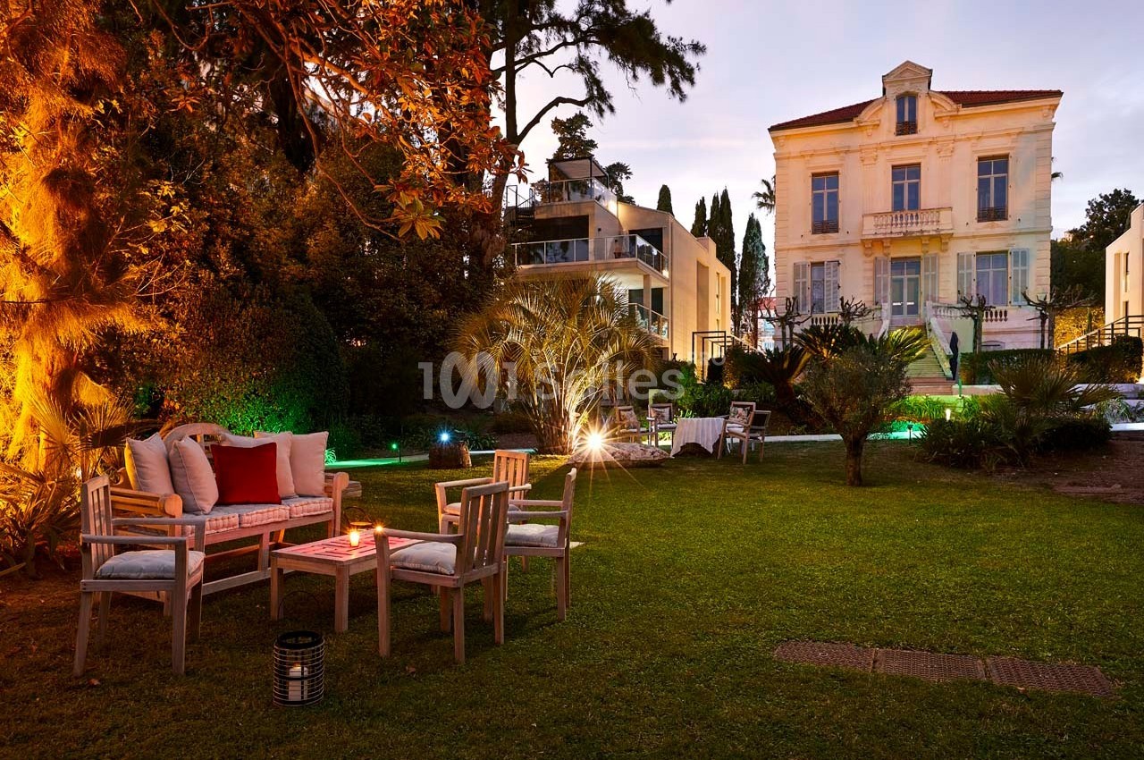 Jardin aménagé avec mobilier en bois, éclairage chaleureux et vue sur une grande maison ancienne au crépuscule.