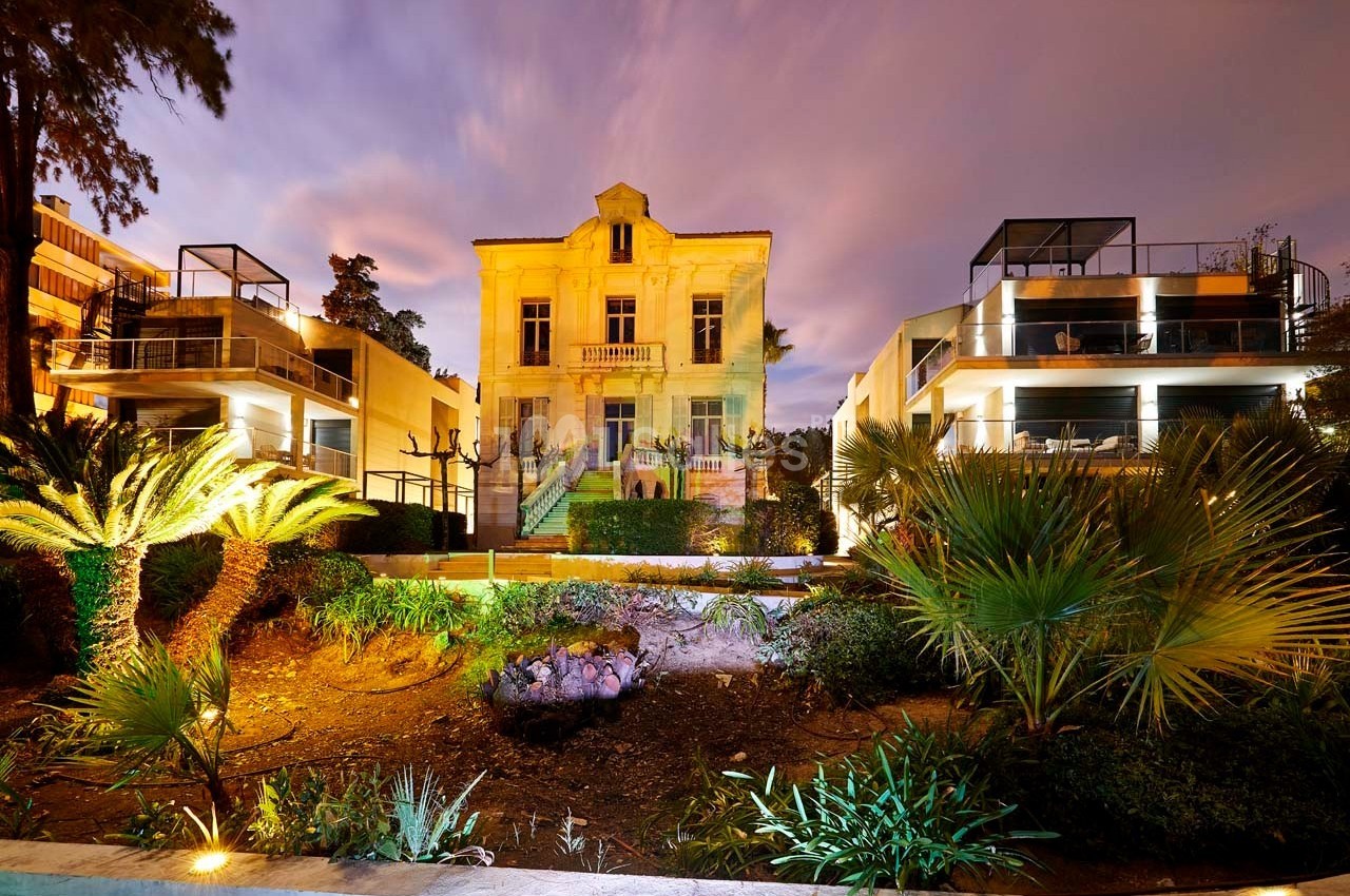 Façade d'une maison ancienne entourée de bâtiments modernes, éclairée la nuit avec un jardin au premier plan.