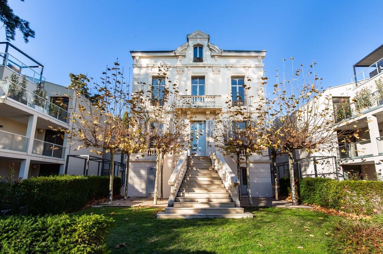 Façade d'un bâtiment blanc à deux étages avec un escalier central, entouré d'arbres et de pelouses.