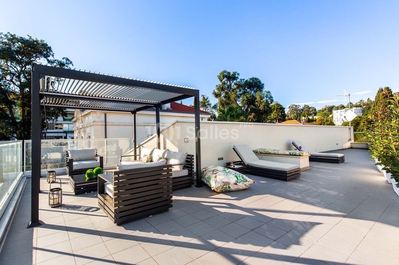 Terrasse spacieuse avec pergola, chaises longues, coussins colorés et vue dégagée sur des arbres et bâtiments voisins.
