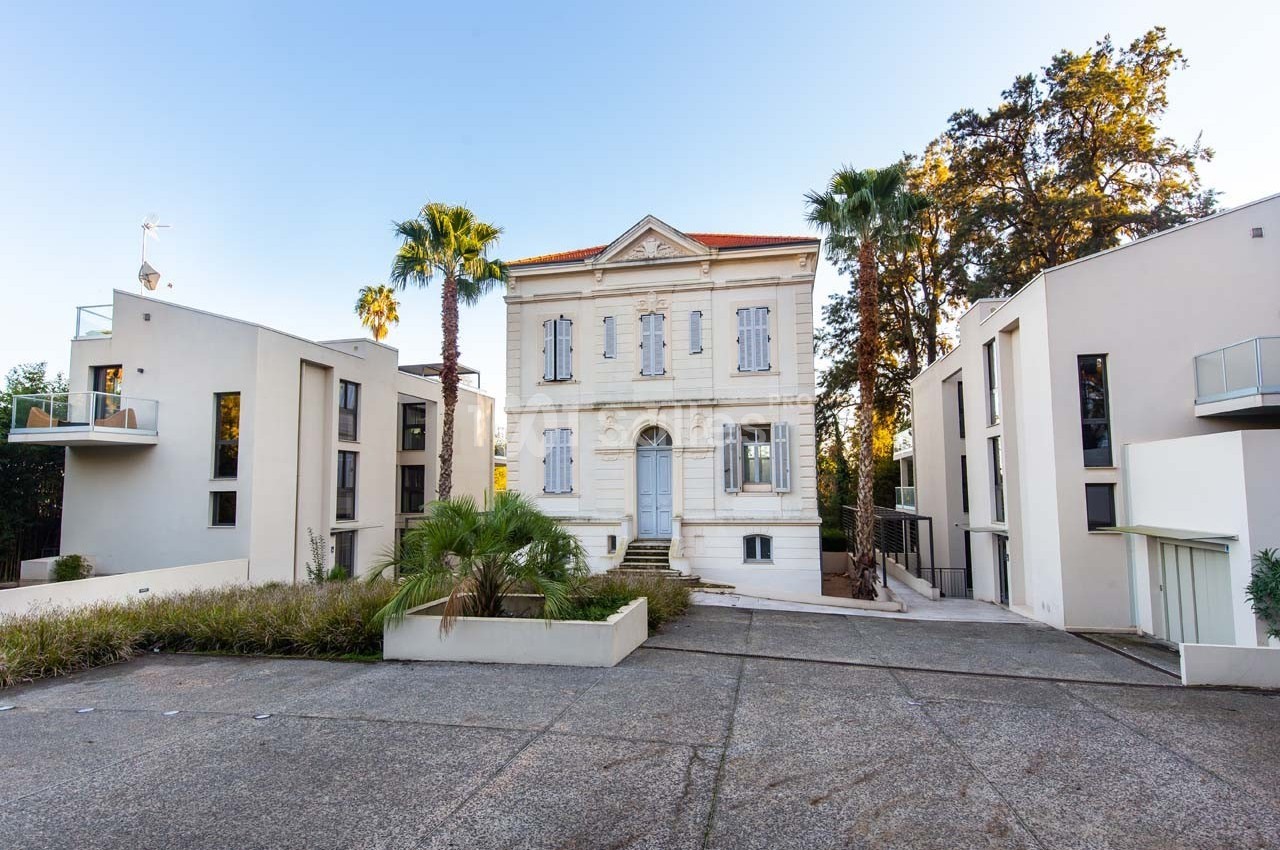 Bâtiment ancien entouré de constructions modernes avec palmiers et allée pavée au premier plan.