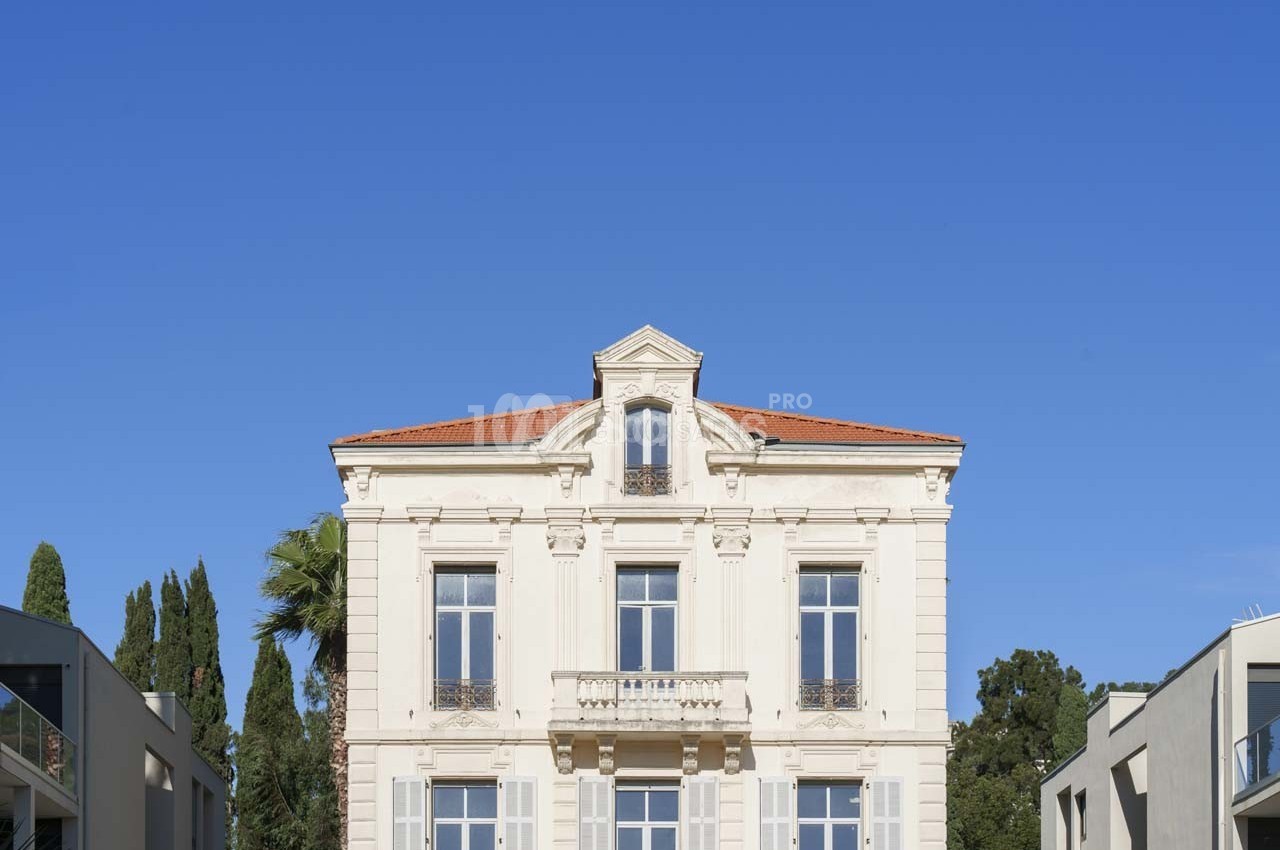 Façade d'un bâtiment ancien à deux étages avec toit en tuiles rouges, entouré d'arbres et de bâtiments modernes.