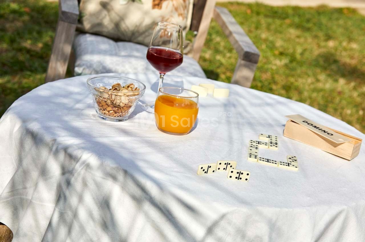 Table ronde en extérieur avec un verre de jus, un verre de vin, des noix, un jeu de dominos et une nappe blanche.