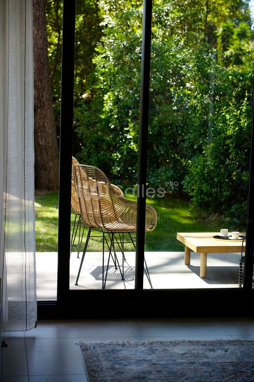 Deux fauteuils en rotin et une table basse avec une tasse, sur une terrasse donnant sur un jardin verdoyant.